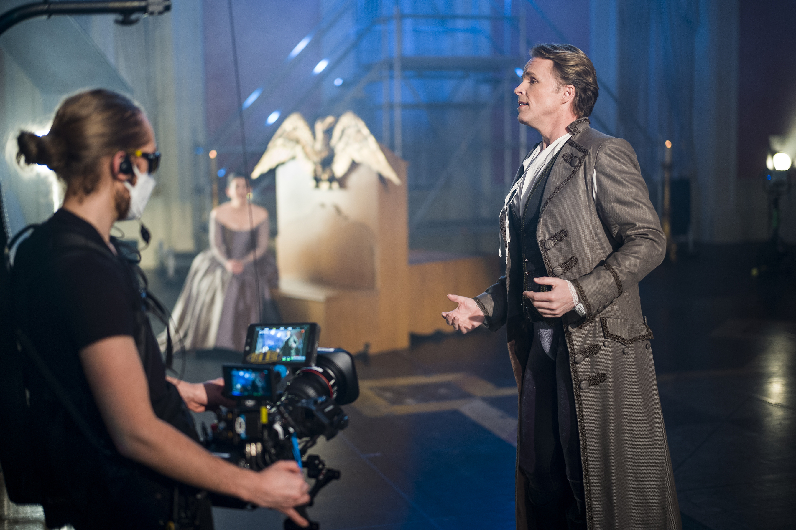 In the foreground a film opperator is shoot a performer wearing a long coat